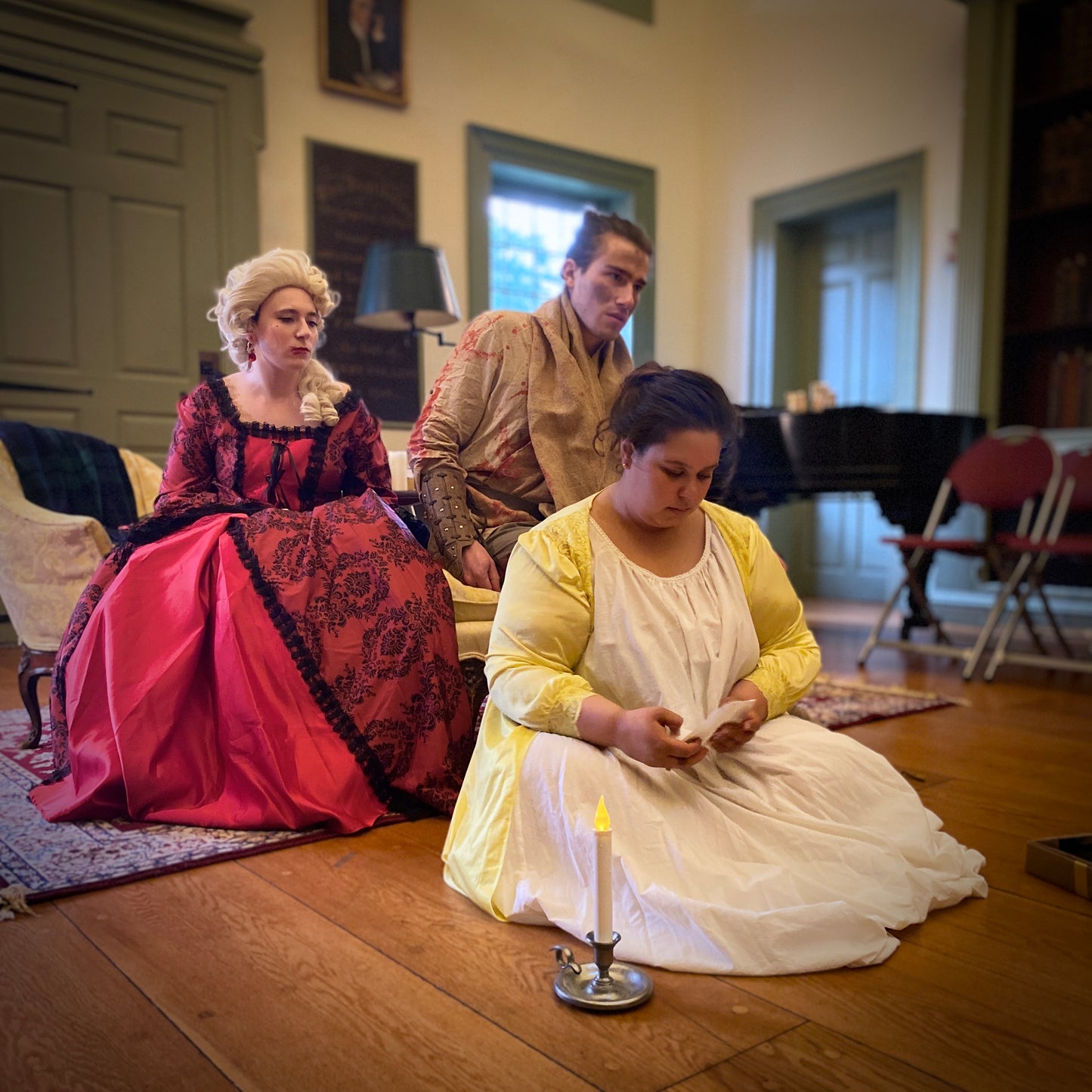 Cast of Valkyrie Productions Pay No Heed in period costumes sitting on the wooden floor in Redwood Library with a candle in a room.