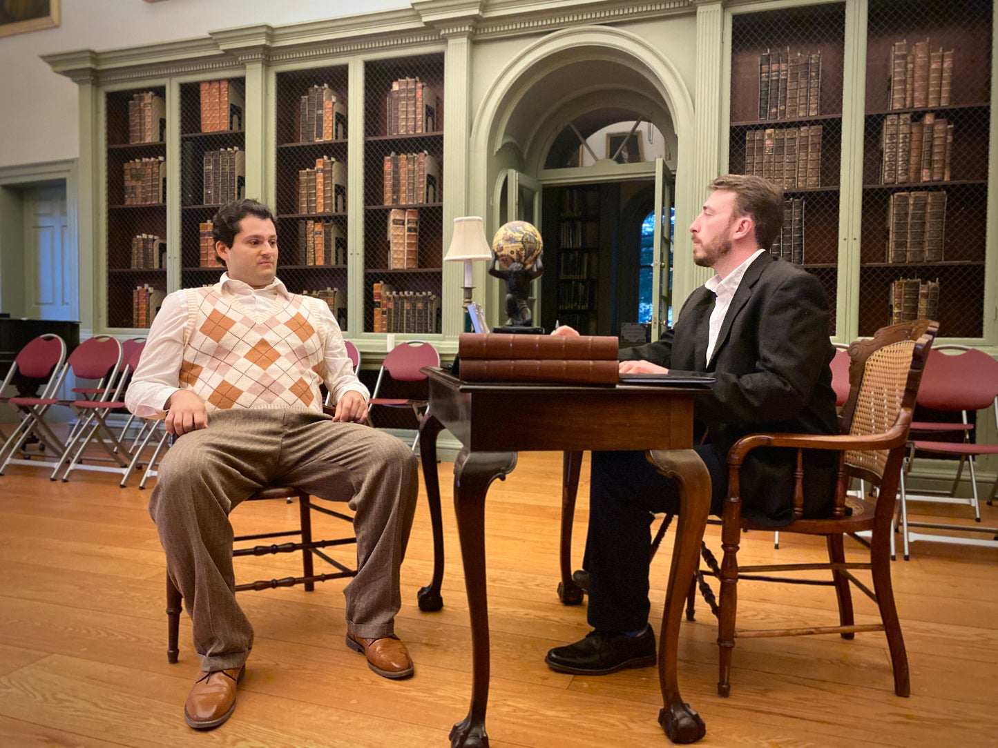 Cast of Valkyrie Productions' Pay No Heed sitting at a table in a library with bookshelves in the background