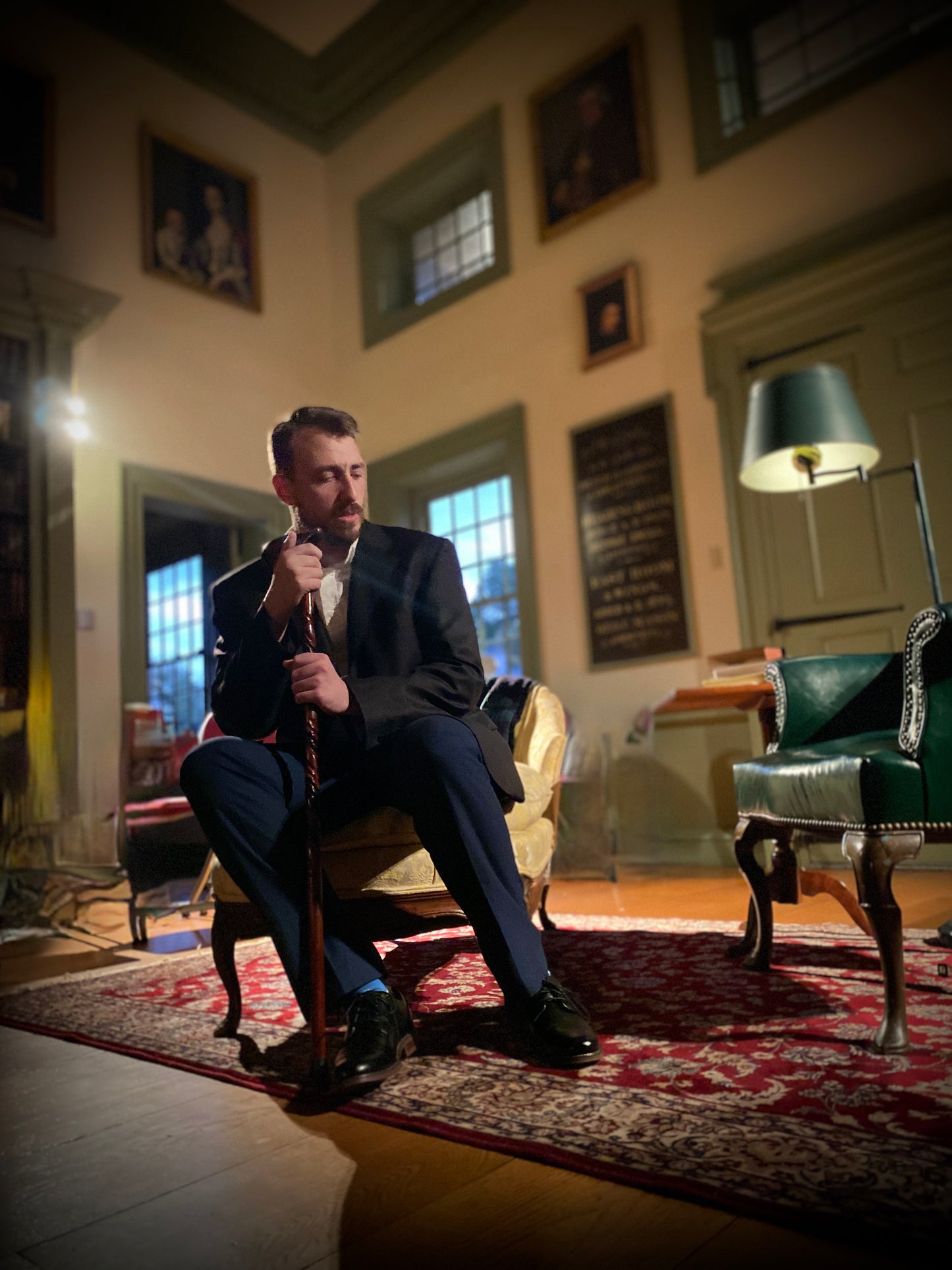The Captain in Valkyrie Productions' Pay No Heed sitting on a yellow chaise in Redwood Library with classical decor, holding a cane.