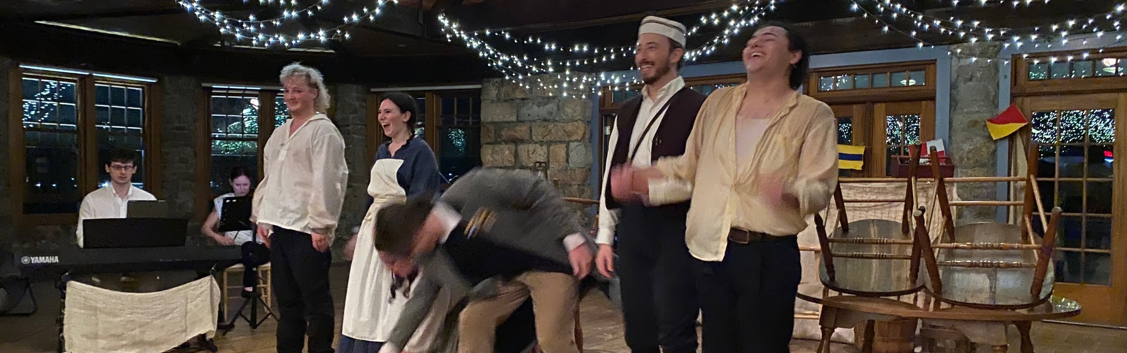 Valkyrie Productions's Sea Fever cast in period costumes in a room with string lights and wooden furniture taking final bow.