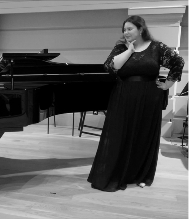 Opera singer Meghan Thomson in formal attire standing next to a grand piano in a room with a window.